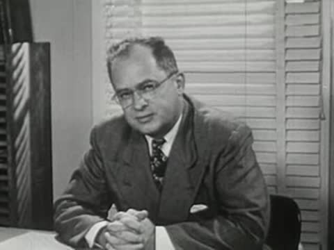 A man in a suit talking at a desk with blinds behind