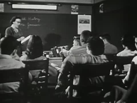 Teacher standing by blackboard in front of seated students in classroom.