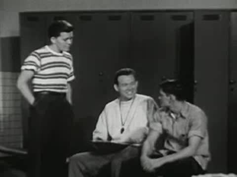 Coach sitting down and talking with students in locker room.