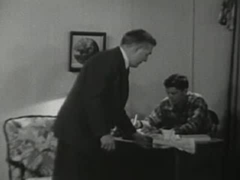 Father walking over to son studying at a desk in the evening.