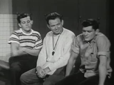 Three young men sitting on a bench, one standing up.