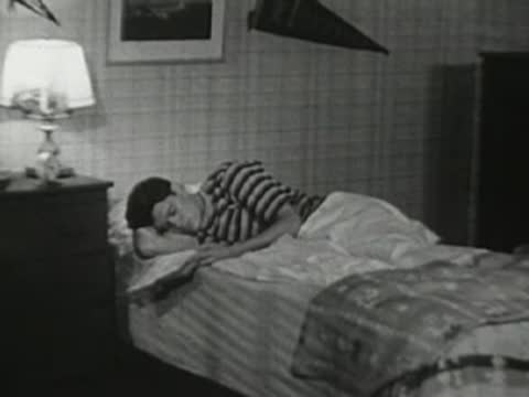 Wide shot of the teenager in bed as he gets up to turn off the alarm.