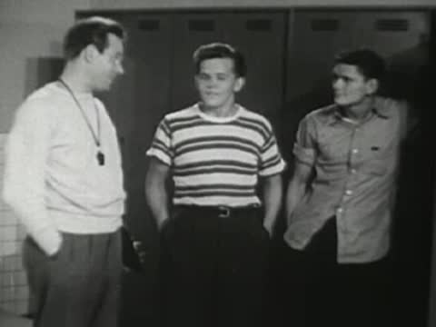 The coach stands talking to two young athletes near lockers.