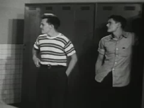Two athletes stand near lockers in a locker room, adjusting their clothing.