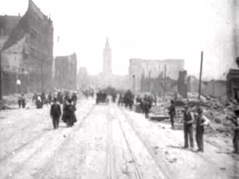 Pedestrians walking towards the camera on a wide debris-strewn avenue.