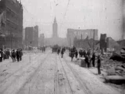 Crowds of pedestrians walking along the ruins of a post-earthquake city street.