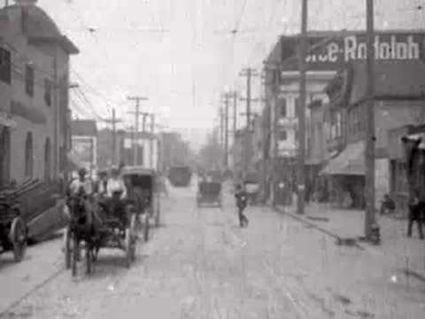 City street featuring storefronts, carriages, and people walking about.