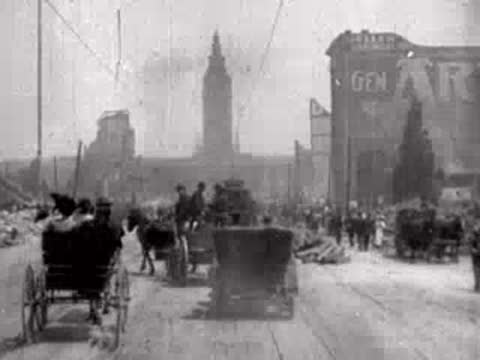 Carriages and pedestrians travel along a wide street flanked by earthquake-damaged buildings.