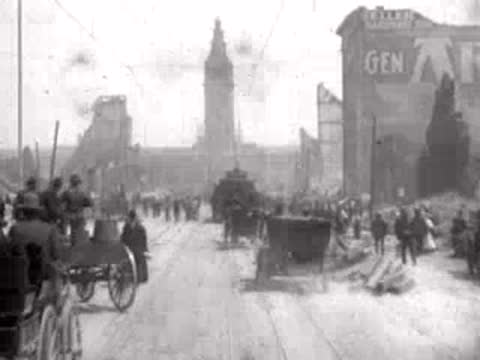 People walk past horse-drawn carriages and ruins on a devastated city street.