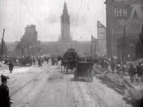 A horse-drawn vehicle travels down a dusty street amid ruins and walking crowds.