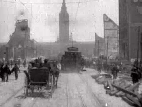 Carriages and pedestrians navigate a wide, dusty street surrounded by urban devastation.