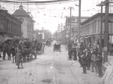 People gathering on a wide city street with horse-drawn carriages.