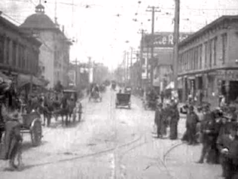 Crowds and carriages move along a city street lined with storefronts.