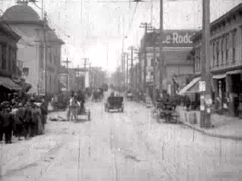Commercial street view with storefronts, pedestrians, and moving traffic.