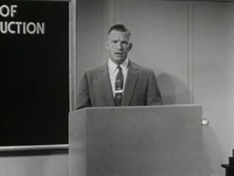 Man standing at podium with text overlay 'OF UCTION' and blackboard