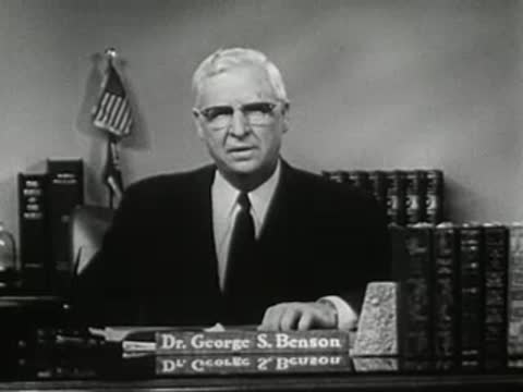 Man speaking behind desk with nameplate 'Dr. George S. Benson' and bookshelf background