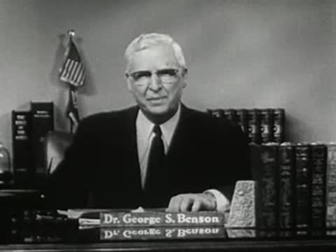 Man speaking behind desk with nameplate 'Dr. George S. Benson' and bookshelf background