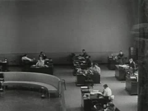pan from a man in a suit speaking to an office workplace scene