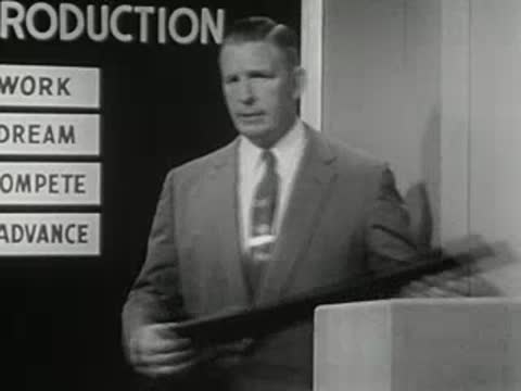 man in a suit gesturing toward text blocks on a set board