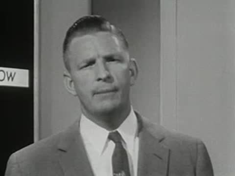 A man in a suit speaks in front of a dark chalkboard background.