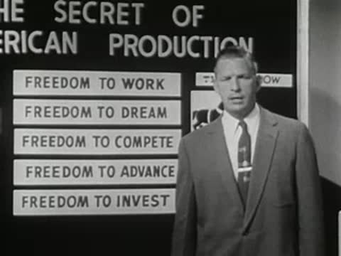 A speaker stands before a chalkboard listing freedoms and production secrets.