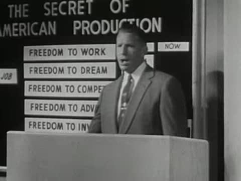 A speaker at a lectern gestures toward a chalkboard with listed freedoms.