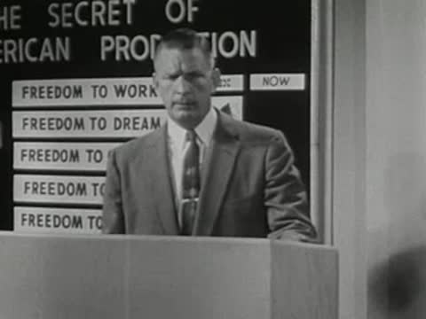 A speaker stands at a lectern delivering a presentation before a chalkboard.