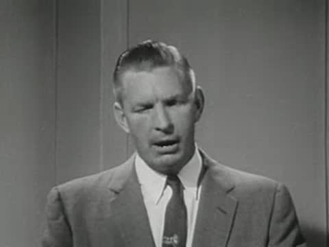 A man in a suit speaks closely to the camera against a plain background.
