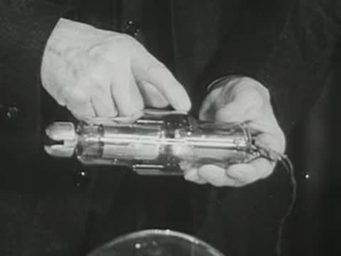Lecturer in suit points to components of an X-ray tube held in hand.