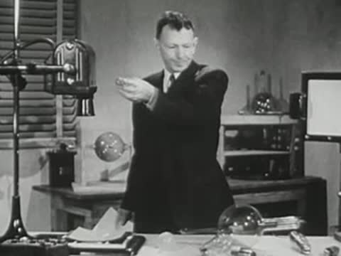 Lecturer holds a small object, gestures, and holds a paper document in hand.