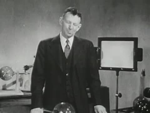 Man in a suit stands in a laboratory, looking down and adjusting equipment.
