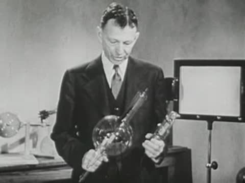 Man in a suit stands in a laboratory, holding glass apparatus.