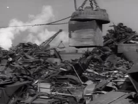 An overhead crane lifting and moving a large cube of scrap metal.