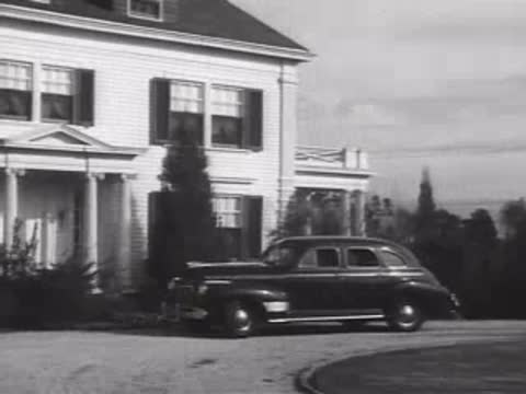 A classic 1940s sedan driving out through brick gate pillars of an estate.