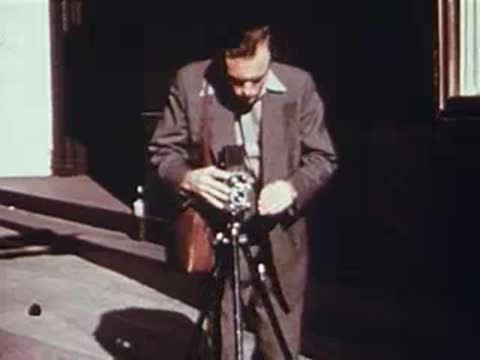 A man in a suit adjusting a vintage camera on a tripod outside.