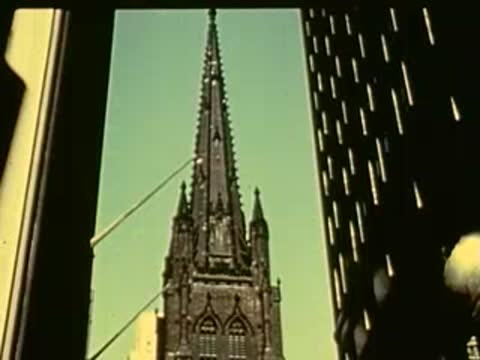 Camera pans down a gothic church steeple between modern buildings.