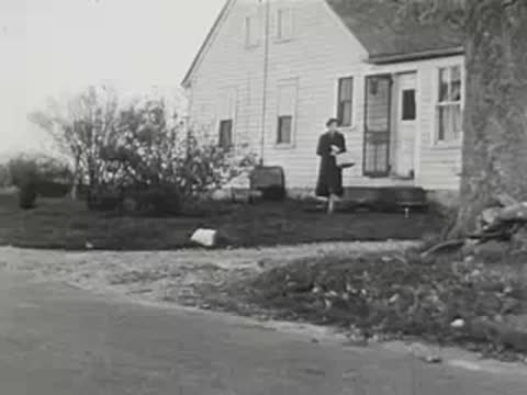 A woman stands outside a white house with a basket, black and white.