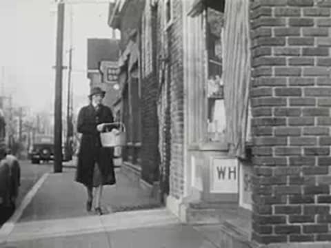 A woman carrying a basket walks down a sidewalk past brick buildings.