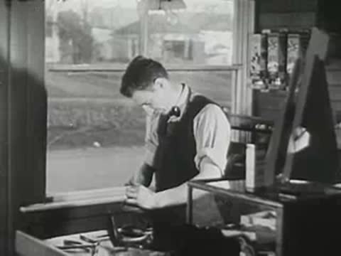 A man works at a counter in a shop, black and white footage.