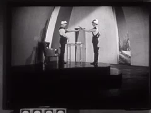 Two people in futuristic uniforms standing near a table with objects in a studio.