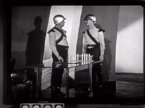 Two people in futuristic uniforms moving around a table in a studio.