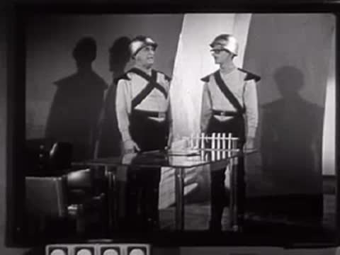 Two people in futuristic uniforms standing near a table with vertical rods.