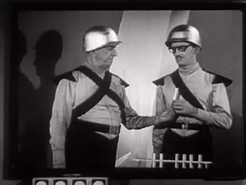 Two people in futuristic uniforms interacting near a table with vertical props.