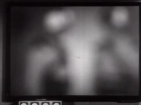 A man looks at a monitor console in a control room, mid-century style.