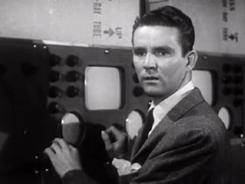 A man operates equipment at a control room console with monitors, mid-century.