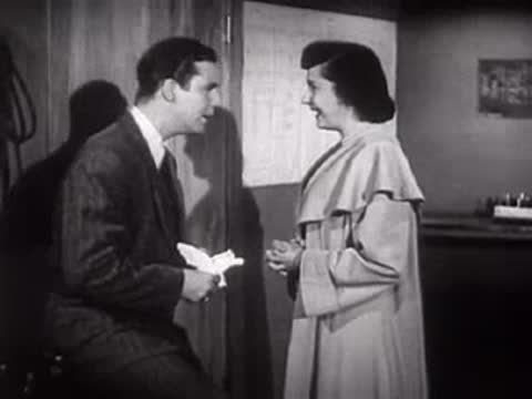 A man and woman stand talking in an office setting, mid-century.