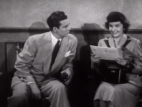 A man and woman sit together looking at a newspaper in a waiting room.