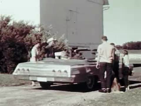 People standing around a convertible car parked outside a building on a farm.