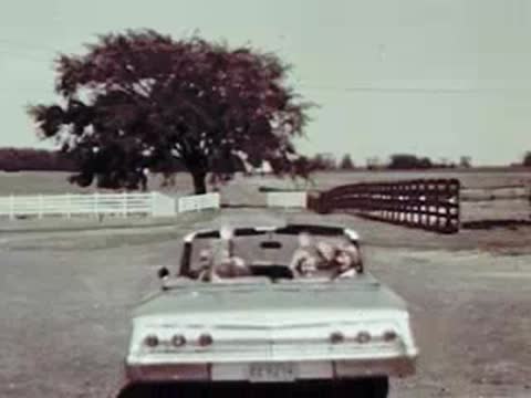 A convertible car driving away down a country road while passengers wave back.