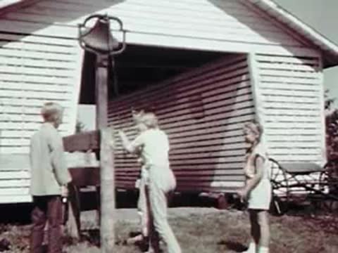 Children standing outside a white barn ringing a large metal bell.